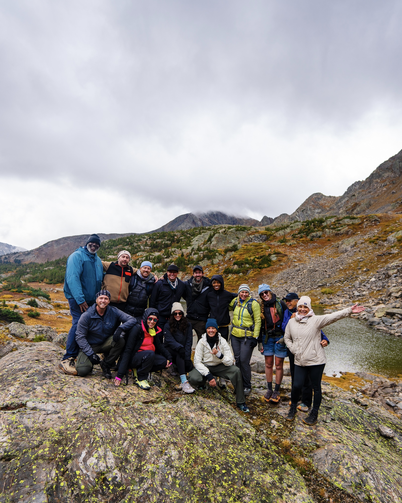 The group smiling together on lichen-covered rocks beside an alpine lake