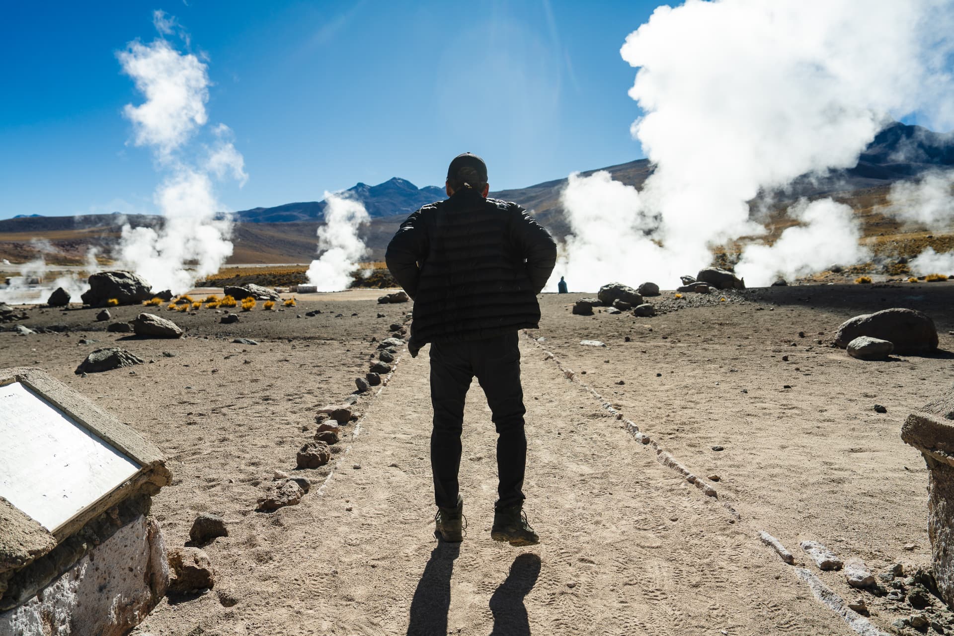 A participant standing before steaming geysers beneath high Andean peaks at El Tatio