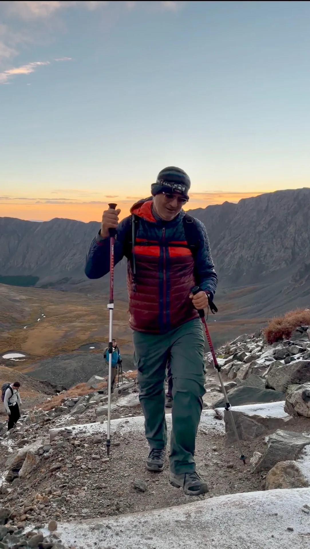 A climber descending a rocky trail on trekking poles at sunrise, alpine valleys below