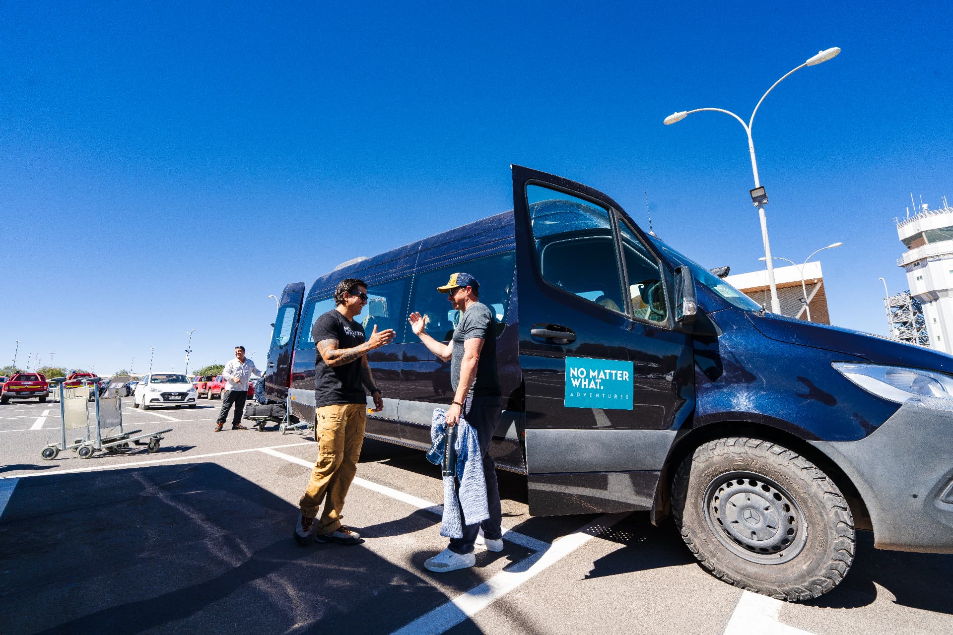 Two men greeting each other beside the No Matter What Adventures van at an airport pickup